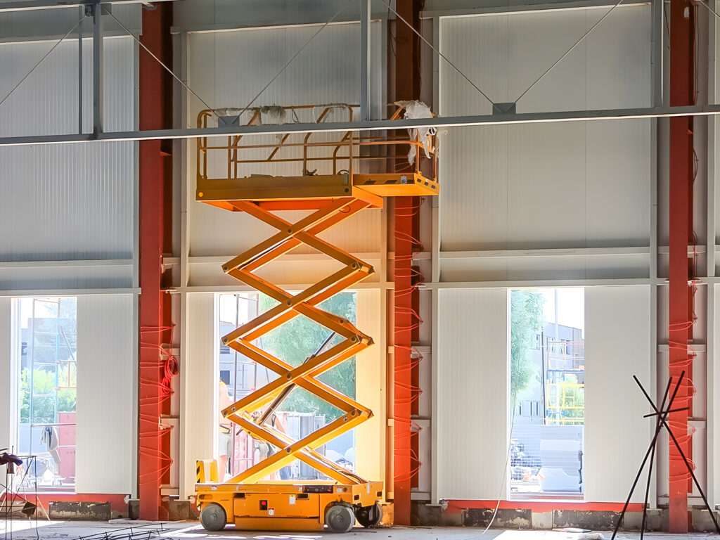 Eine orangefarbene Scherenhebebühne steht in einem im Bau befindlichen Gebäude in der Nähe von großen Fenstern und Stahlträgern.